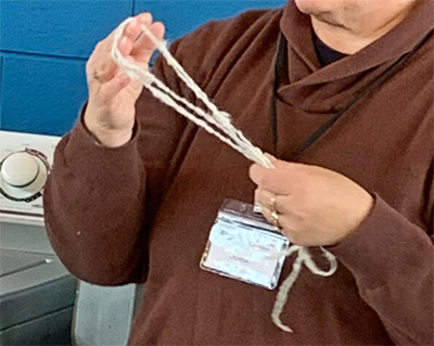 Donna's card challenged her to spin a botanical fiber (flax, hemp, nettle, rose fiber, etc.) and create a texture in the yarn without adding new fiber. On the left she is holding pineapple fiber yarn she spun and on the right, rose fiber or hemp yarn. She concluded that flax and hemp yarns were relatively rough and easy to add texture to while rose fiber was very slippery and fine, making it hard to add texture to. Pineapple fiber was intermediate, soft and smooth and able to hold texture. She was most impressed with the texture and interest of yarn in which she spun all the fibers together.