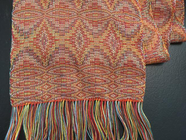 From Shelby: I added an additional two yards of warp and wove this scarf at the end of class. I had a four-color warp in yellow, red, turquoise and dark green, and the weft was orange. This was an Echo pattern, but I combined a couple of the drafts that we were supplied with to add the lozenges at the border of the scarf.